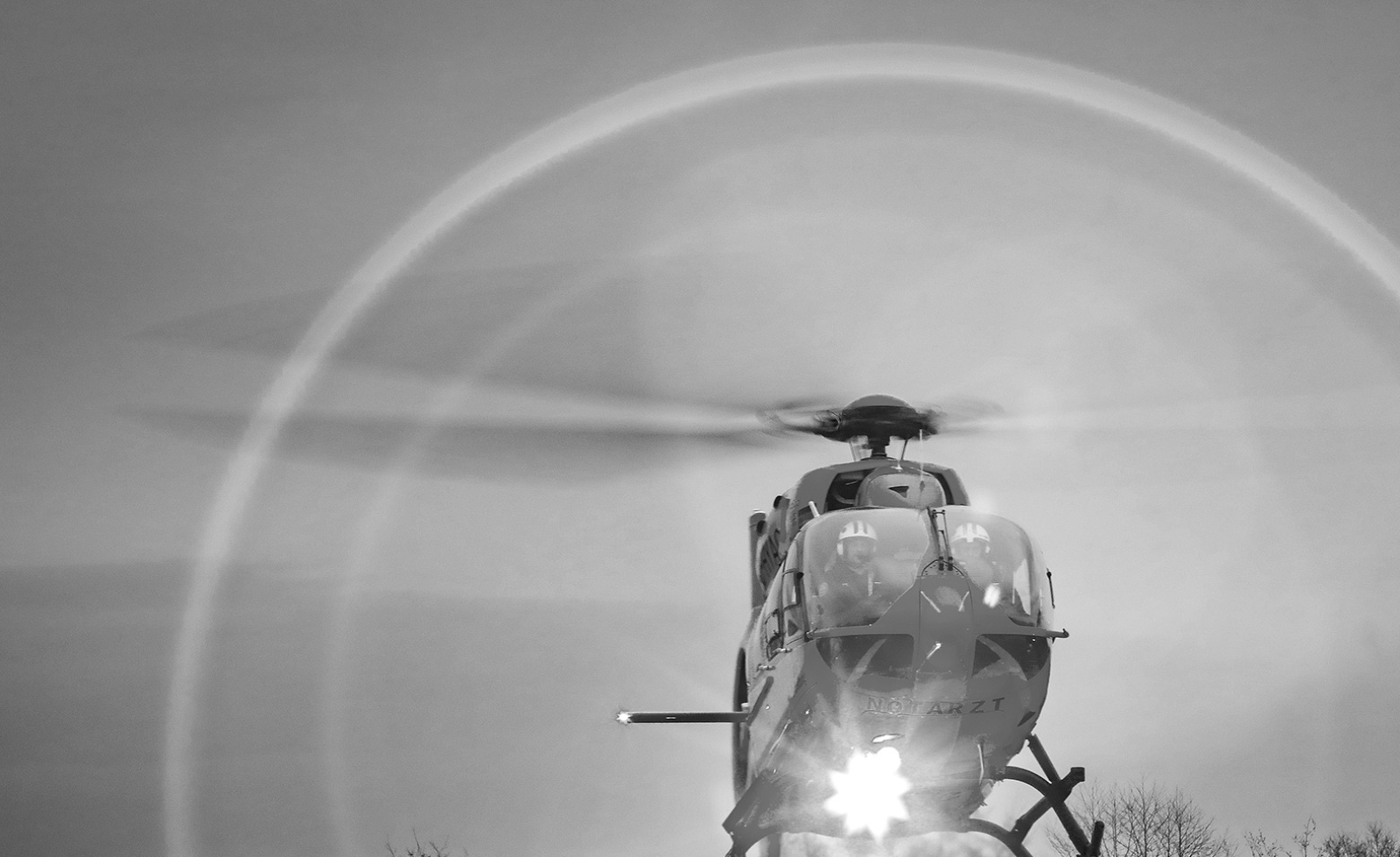 Rettungshubschrauber frontal beim Start, Rotorkreis sichtbar, zwei Piloten mit Helmen im Cockpit.