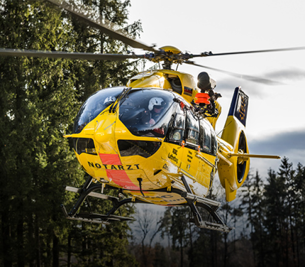 ADAC Luftrettung Rettungshubschrauber mit Besatzung im Tiefflug über Wald, Pilot und Crew an der Tür sichtbar.