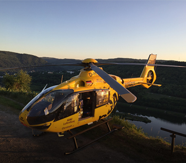 ADAC Luftrettung Rettungshubschrauber gelandet an Flussufer bei Sonnenuntergang, geöffnete Seitenklappe und Besatzung an Bord.