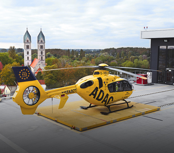 Gelber Rettungshubschrauber der ADAC Luftrettung auf Dachlandeplatz eines Gebäudes, Kirchtürme im Hintergrund.