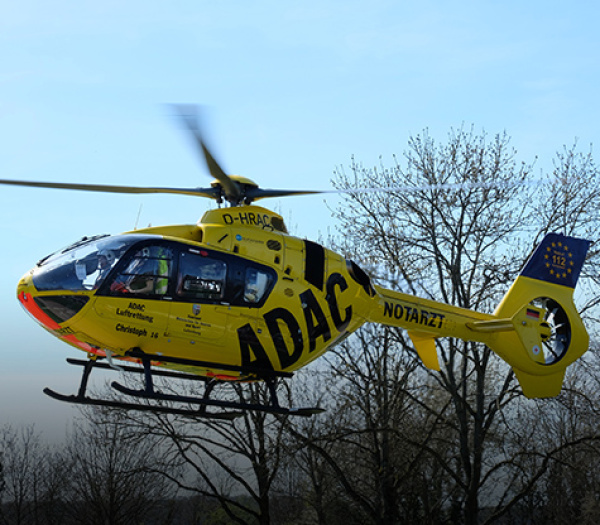 Gelber Rettungshubschrauber der ADAC Luftrettung mit NOTARZT-Beschriftung im Tiefflug vor kahlen Bäumen.