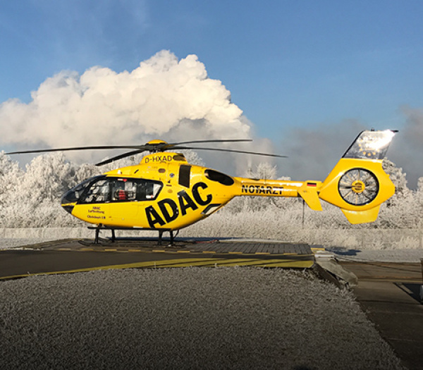 ADAC Luftrettung Rettungshubschrauber auf vereistem Landeplatz vor verschneiter Baumlandschaft.