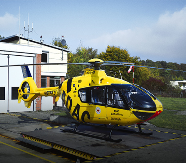Gelber ADAC Luftrettung Rettungshubschrauber auf Landeplatz vor Einsatzgebäude, Beschriftung ADAC sichtbar.