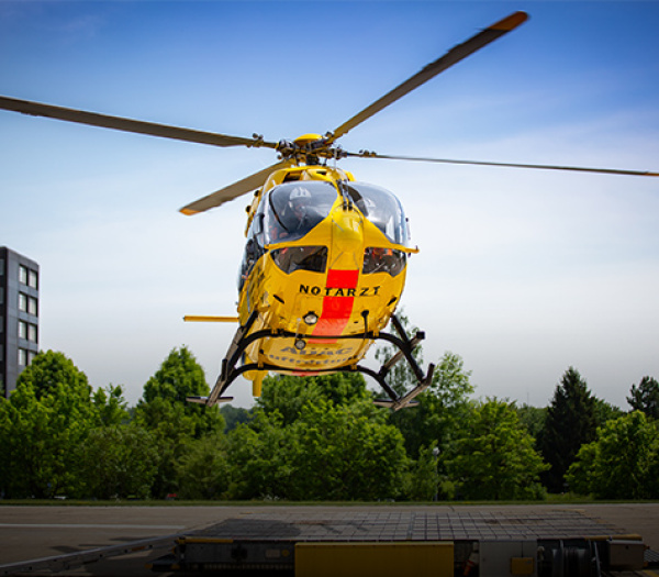 ADAC Luftrettung Rettungshubschrauber mit 'Notarzt'-Schriftzug schwebt über Landeplatz vor Bäumen.