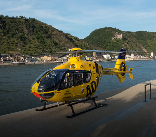 Gelber Rettungshubschrauber der ADAC Luftrettung auf Uferpromenade am Rhein, Hügel mit Burg im Hintergrund.