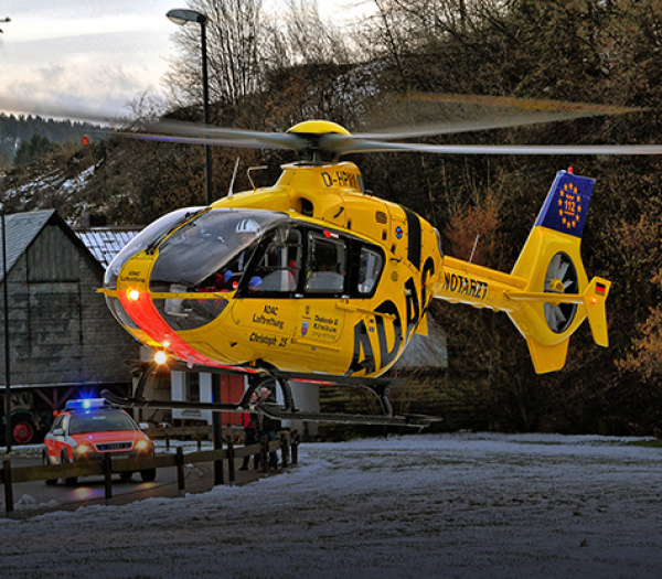Rettungshubschrauber der ADAC Luftrettung landet an verschneiter Landstraße, Rettungswagen mit Blaulicht daneben.