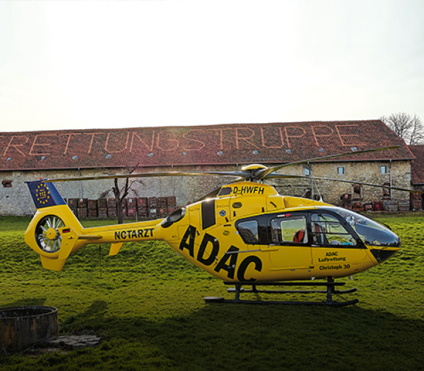 Gelber Rettungshubschrauber der ADAC Luftrettung auf Wiese vor Scheune mit Schrift RETTUNGSTRUPPE.