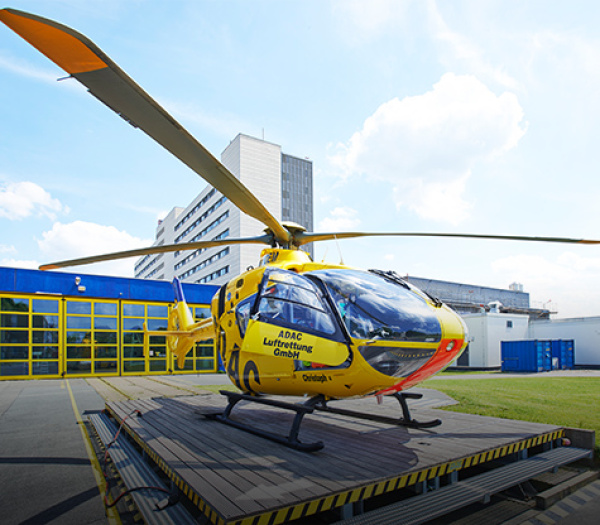 Gelber Rettungshubschrauber der ADAC Luftrettung auf Landeplattform vor Krankenhaus- und Hangargebäuden.