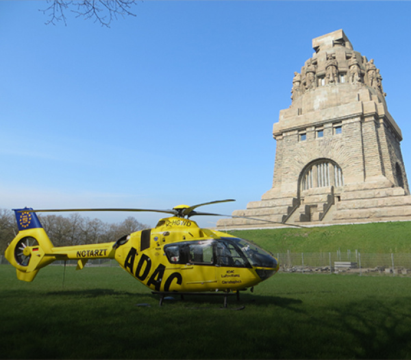 Gelber Rettungshubschrauber der ADAC Luftrettung steht auf einer Wiese vor großem steinernem Denkmal.