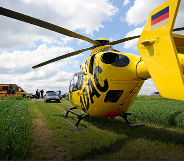 Gelber Rettungshubschrauber der ADAC Luftrettung auf Feld bei Einsatz; Einsatzkräfte und Rettungswagen im Hintergrund.