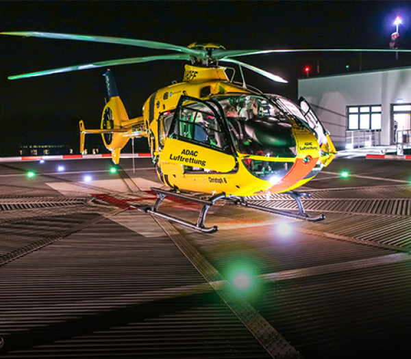 ADAC Luftrettung-Rettungshubschrauber auf beleuchtetem Dachlandeplatz bei Nacht, Cockpit mit Crew sichtbar.