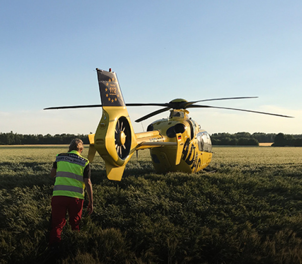 Gelber Rettungshubschrauber der ADAC Luftrettung landet auf Feld, Einsatzkraft in Warnweste geht zum Heck.