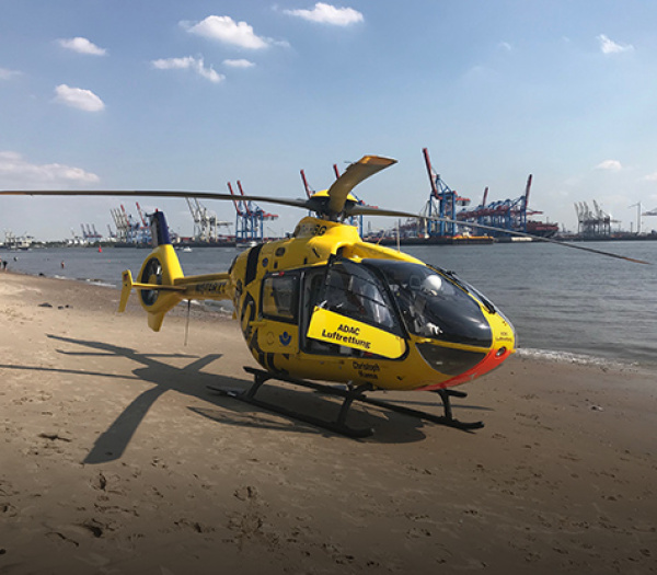 Gelber ADAC Luftrettung Rettungshubschrauber auf Sandstrand am Hafen, Kräne und Wasser im Hintergrund.