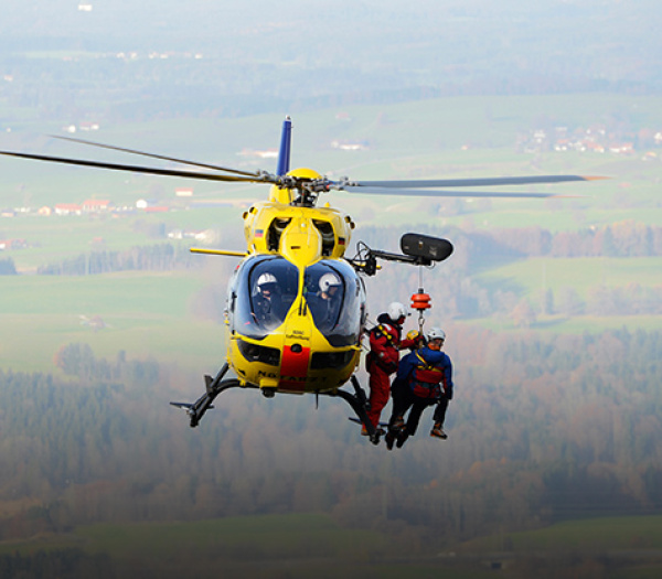 Rettungshubschrauber der ADAC Luftrettung wincht zwei Einsatzkräfte über ländliche Gegend.