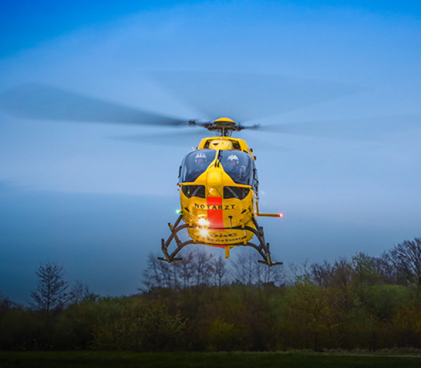 Gelber Rettungshubschrauber der ADAC Luftrettung mit sichtbarer Besatzung schwebt über Waldgebiet bei Dämmerung.