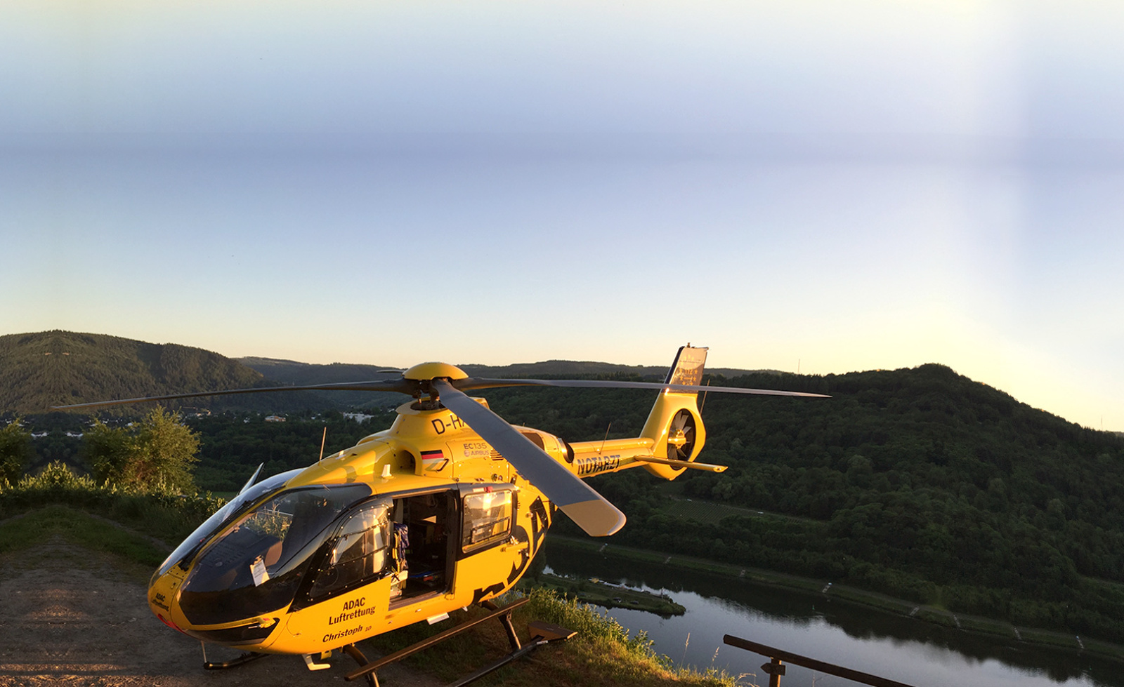 Gelber Rettungshubschrauber der ADAC Luftrettung auf Hanglandeplatz über Fluss und bewaldeten Hügeln bei Abendlicht.