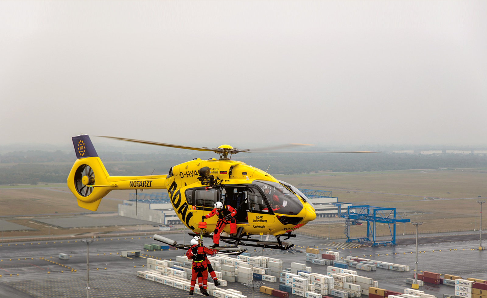 Rettungshubschrauber der ADAC Luftrettung über Container-Terminal, Team seilt zwei Einsatzkräfte per Rettungsseil ab.