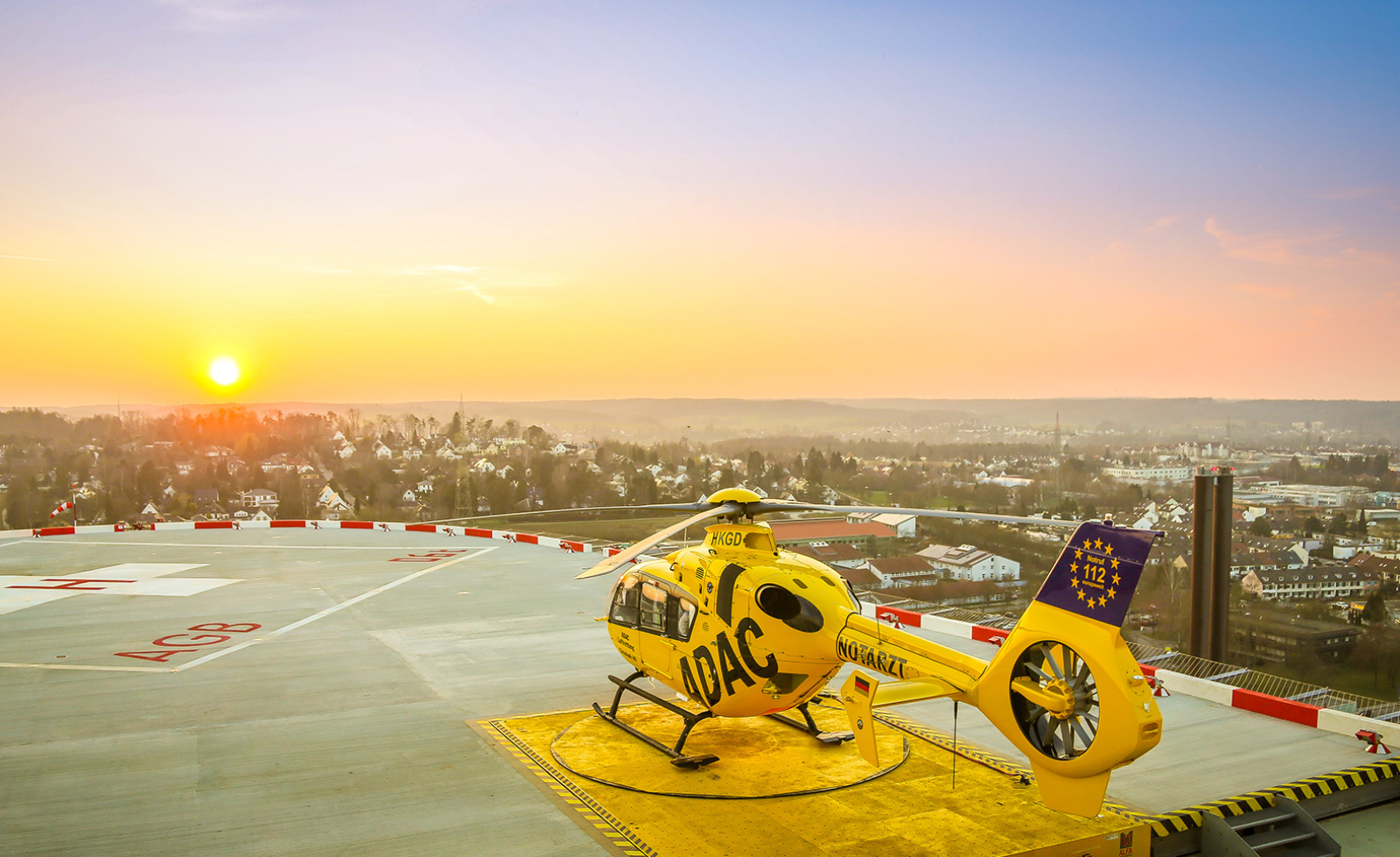 Gelber Rettungshubschrauber der ADAC Luftrettung auf Krankenhaus-Heliport bei Sonnenaufgang über der Stadt