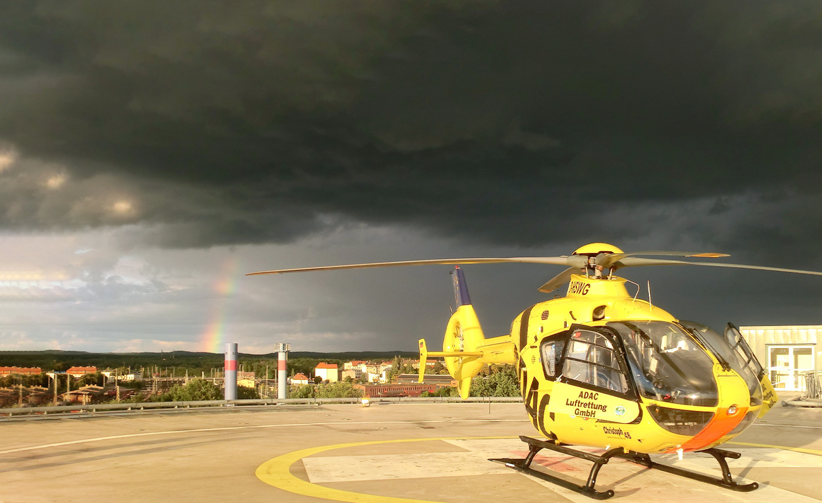 Gelber Rettungshubschrauber der ADAC Luftrettung auf Landeplatz vor dunklen Gewitterwolken mit Regenbogen im Hintergrund.