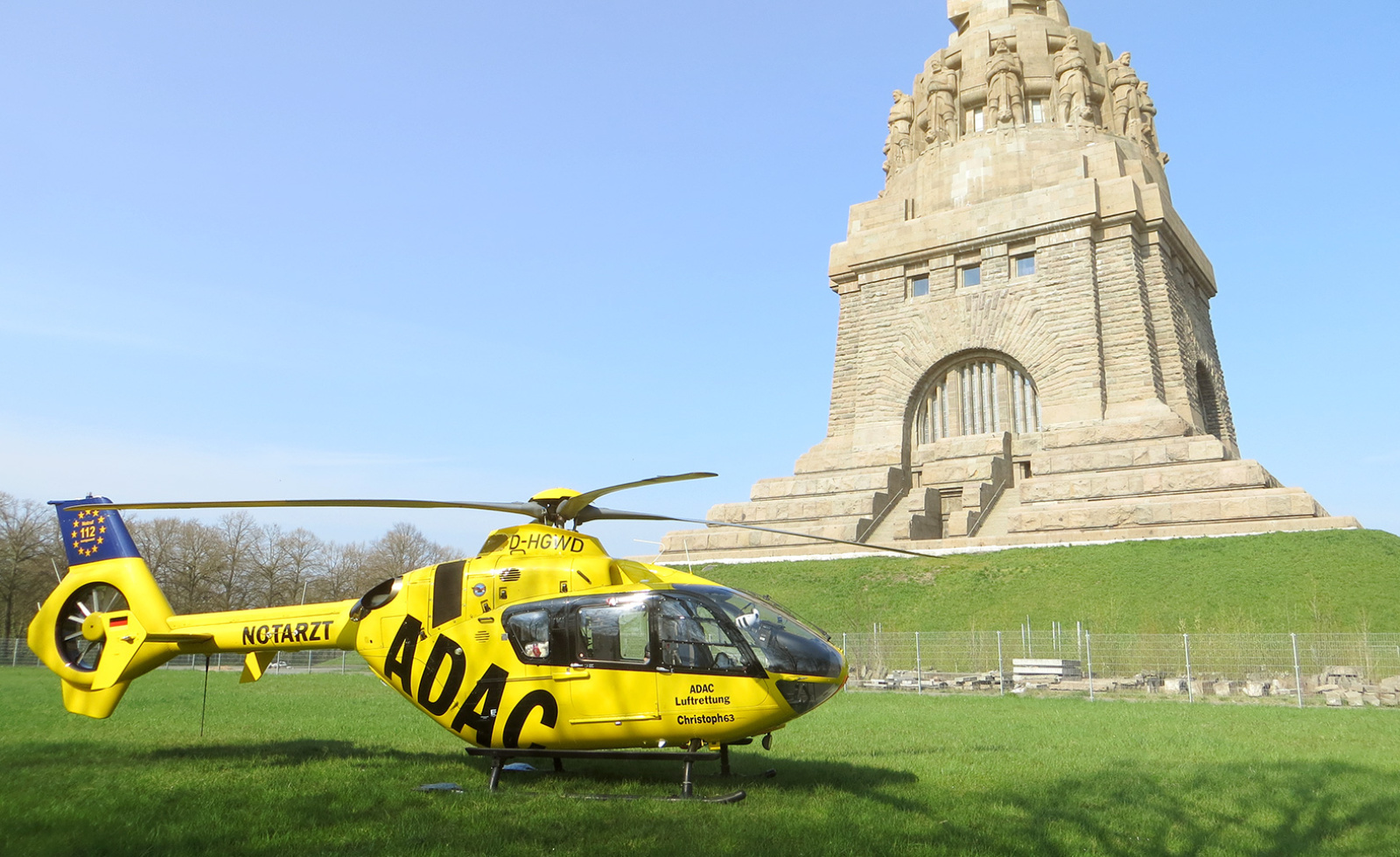 Gelber Rettungshubschrauber der ADAC Luftrettung auf Wiese vor großem steinernen Denkmal.