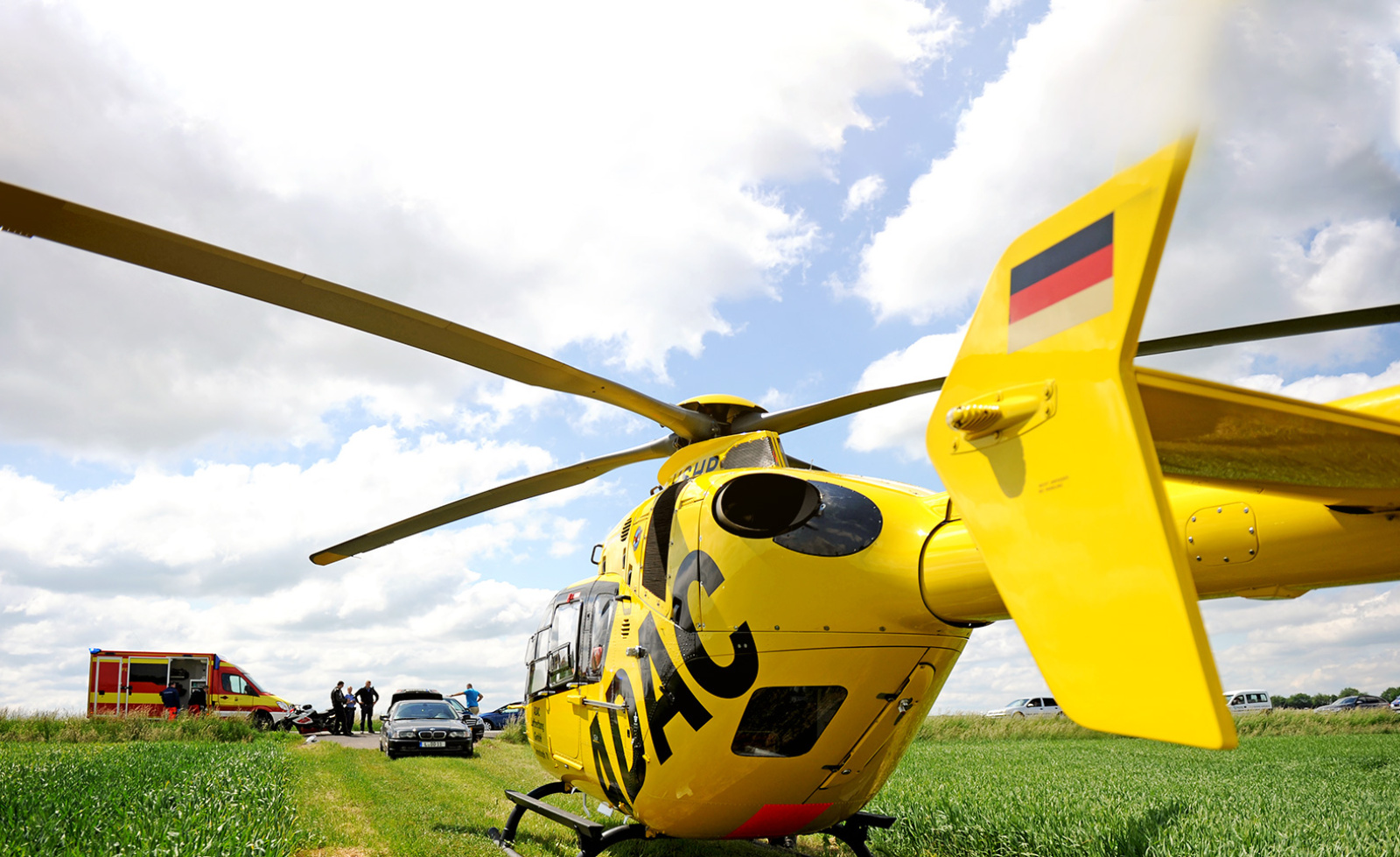 Gelber Rettungshubschrauber der ADAC Luftrettung auf Feld, Einsatzkräfte und Rettungswagen im Hintergrund.