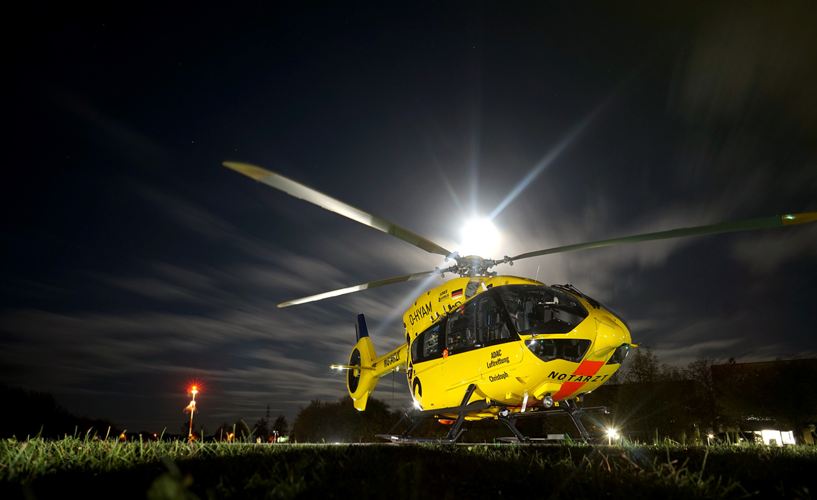 Gelber Rettungshubschrauber der ADAC Luftrettung steht nachts auf Wiese, Rotorblätter vor hellem Mond.