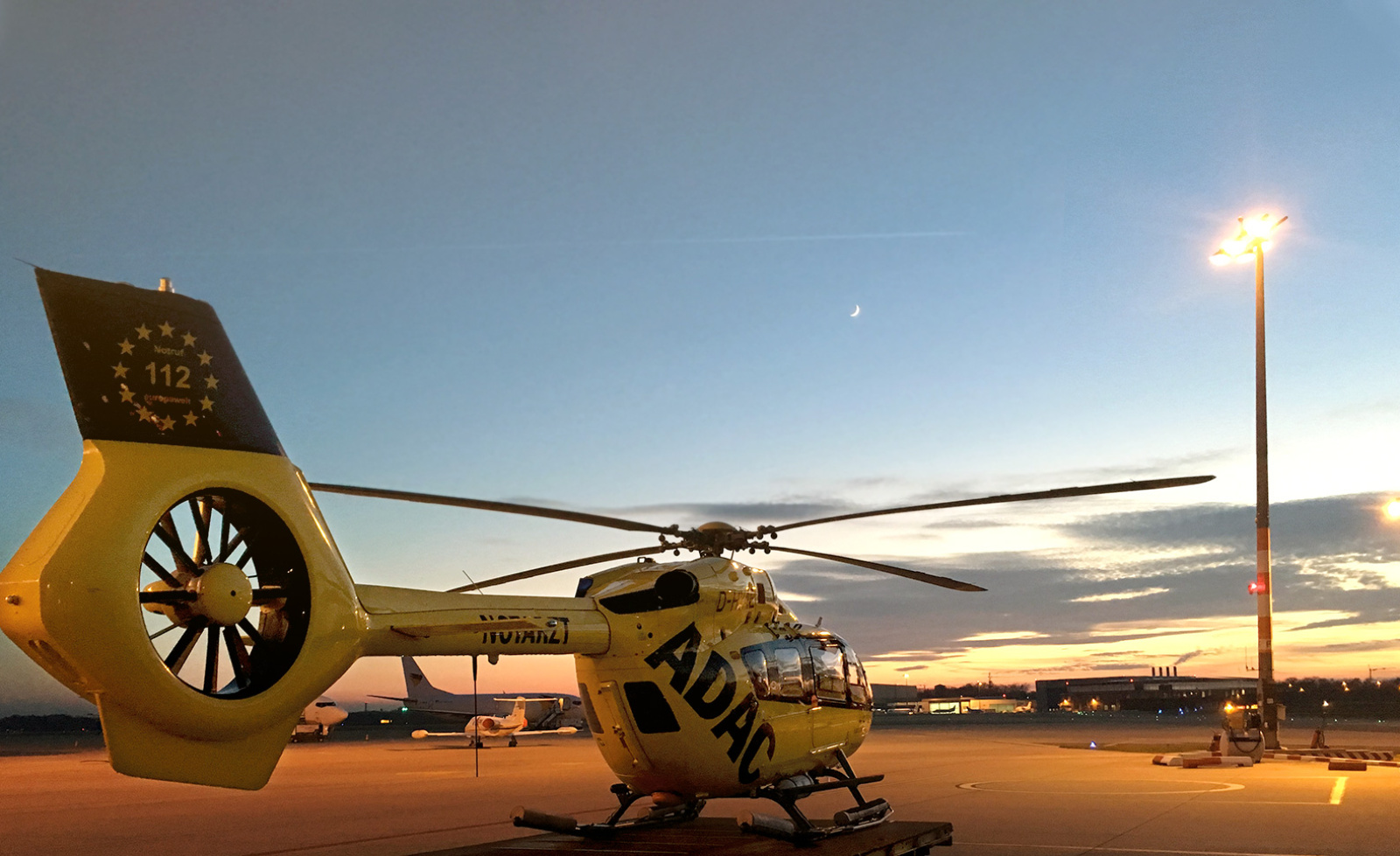 Gelber Rettungshubschrauber der ADAC Luftrettung auf Rollfeld bei Sonnenuntergang, Heck mit Notruf 112 sichtbar.