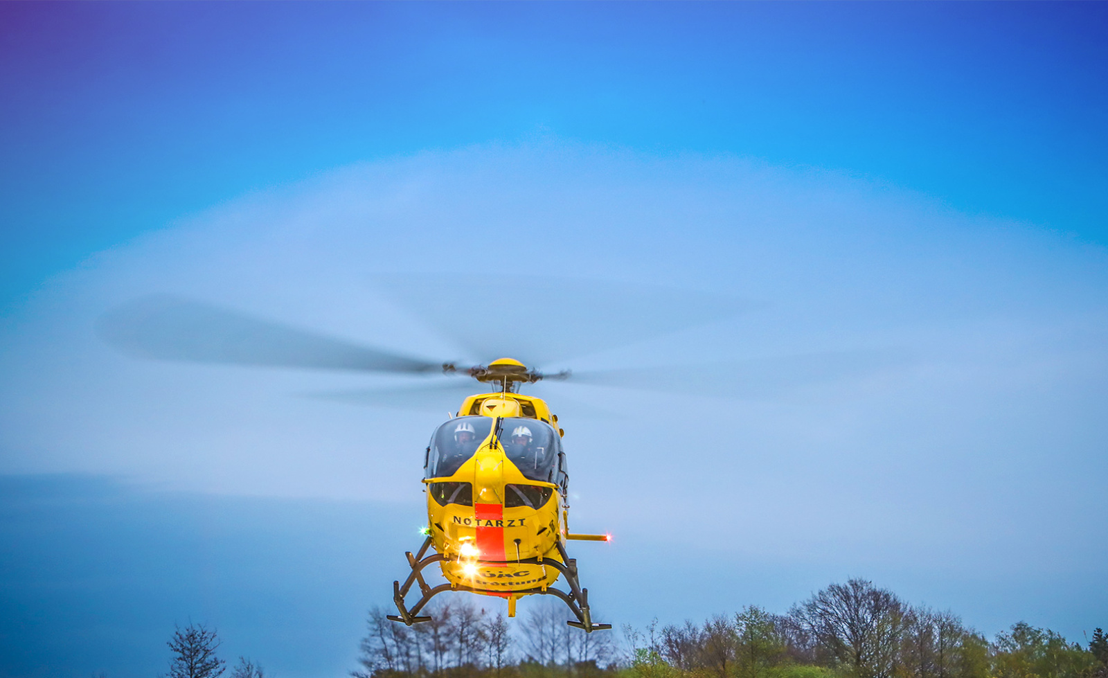 Gelber Rettungshubschrauber mit NOTARZT-Schriftzug und Besatzung im Cockpit schwebt über Baumkrone.
