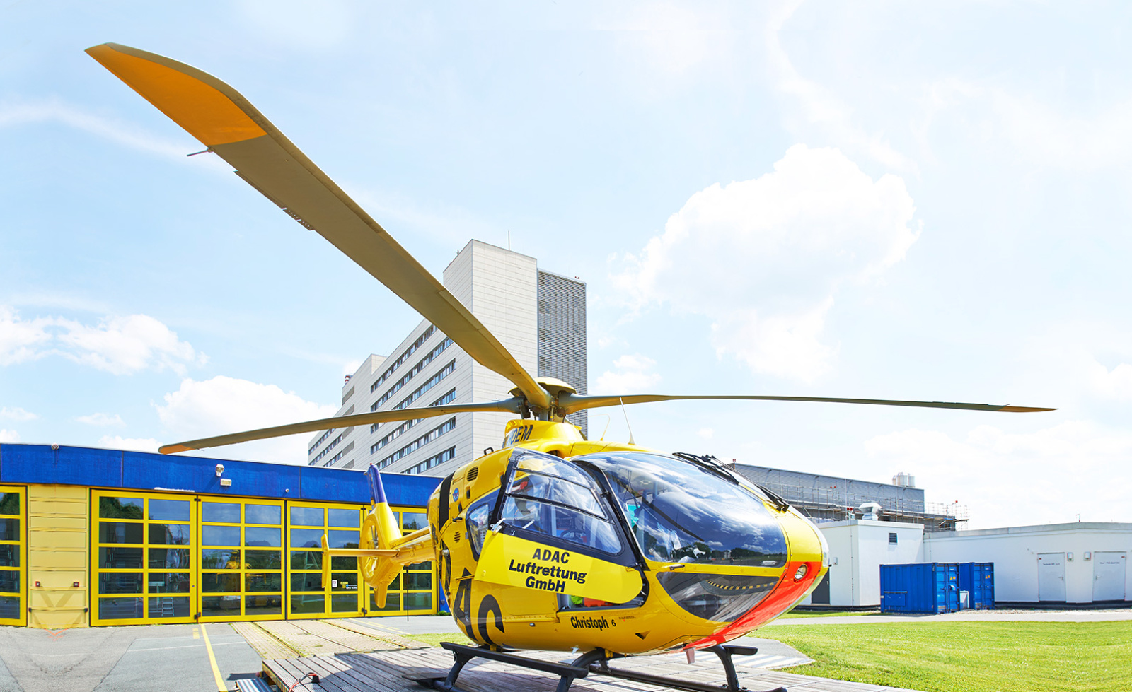 Gelber Rettungshubschrauber der ADAC Luftrettung auf Landeplatz vor Stations- und Hangargebäude.