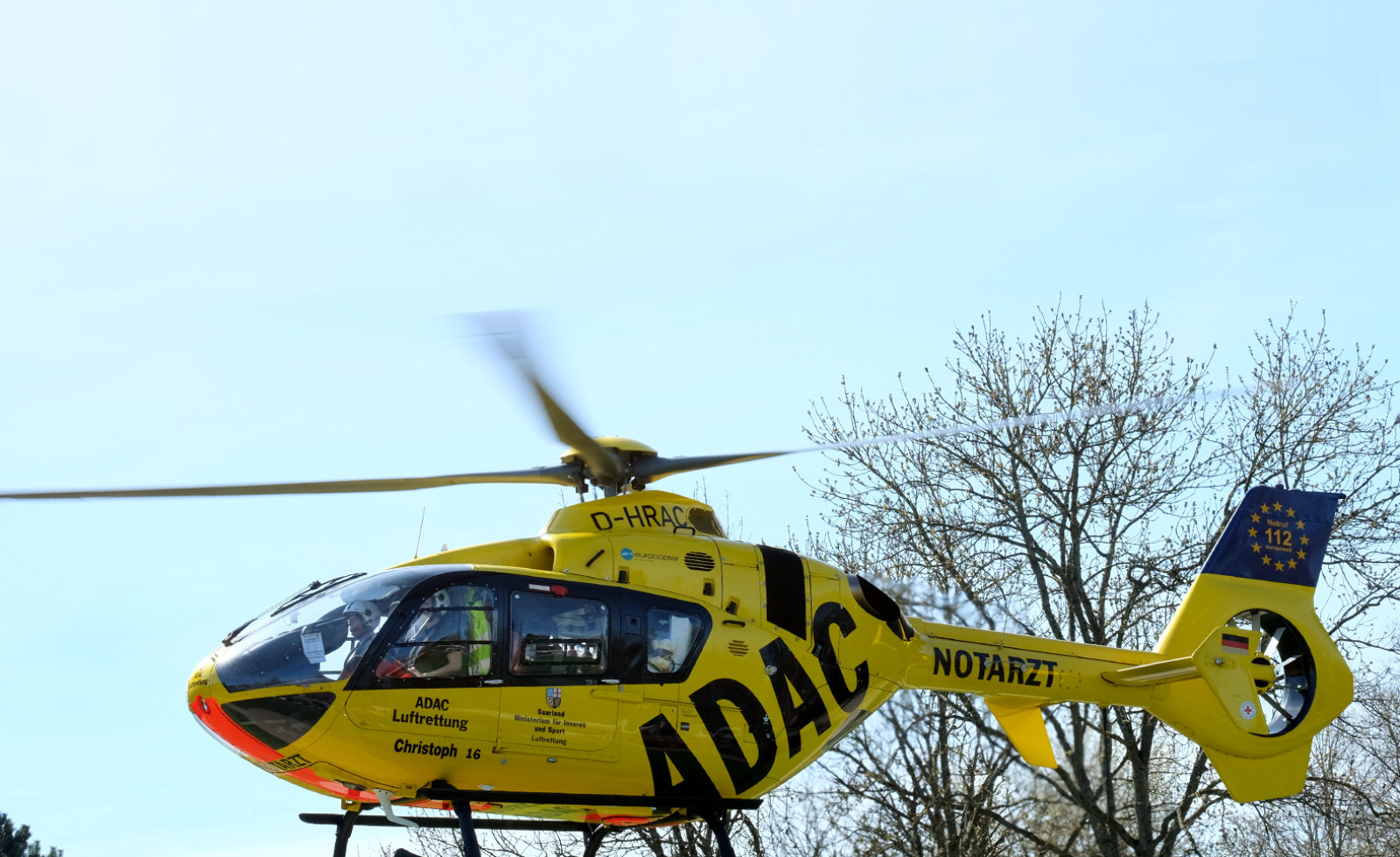 Gelber Rettungshubschrauber der ADAC Luftrettung über Bäumen, Besatzung im Cockpit sichtbar.