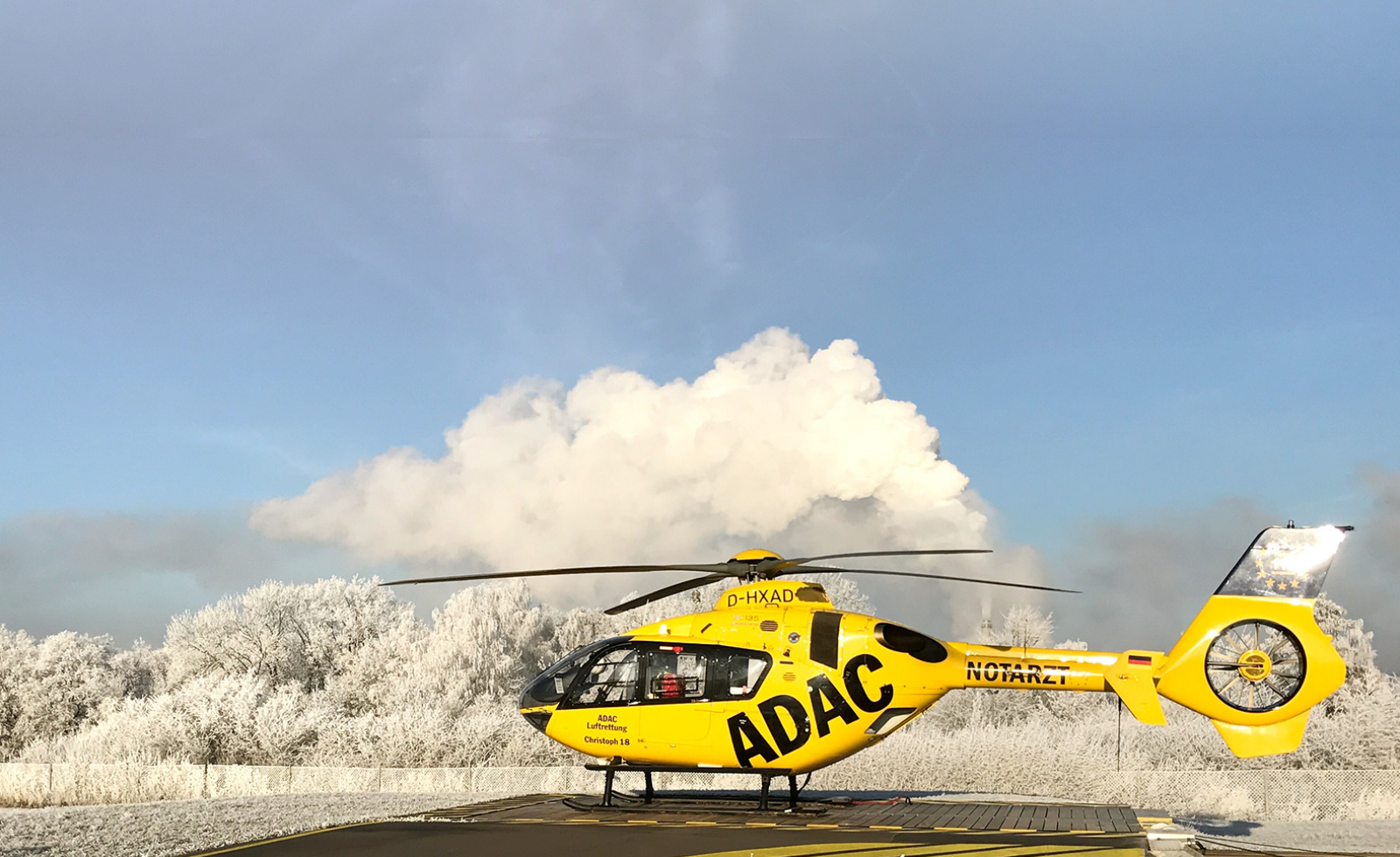 Gelber Rettungshubschrauber der ADAC Luftrettung auf Landeplatz, eingerahmt von frostbedeckten Bäumen und klarem Himmel