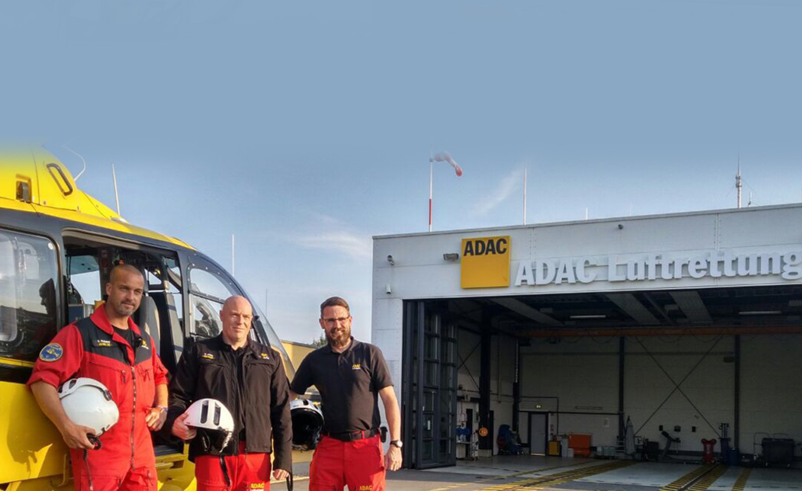 Drei ADAC Luftrettung-Crewmitglieder in Einsatzkleidung vor gelbem Rettungshubschrauber und Hangar.