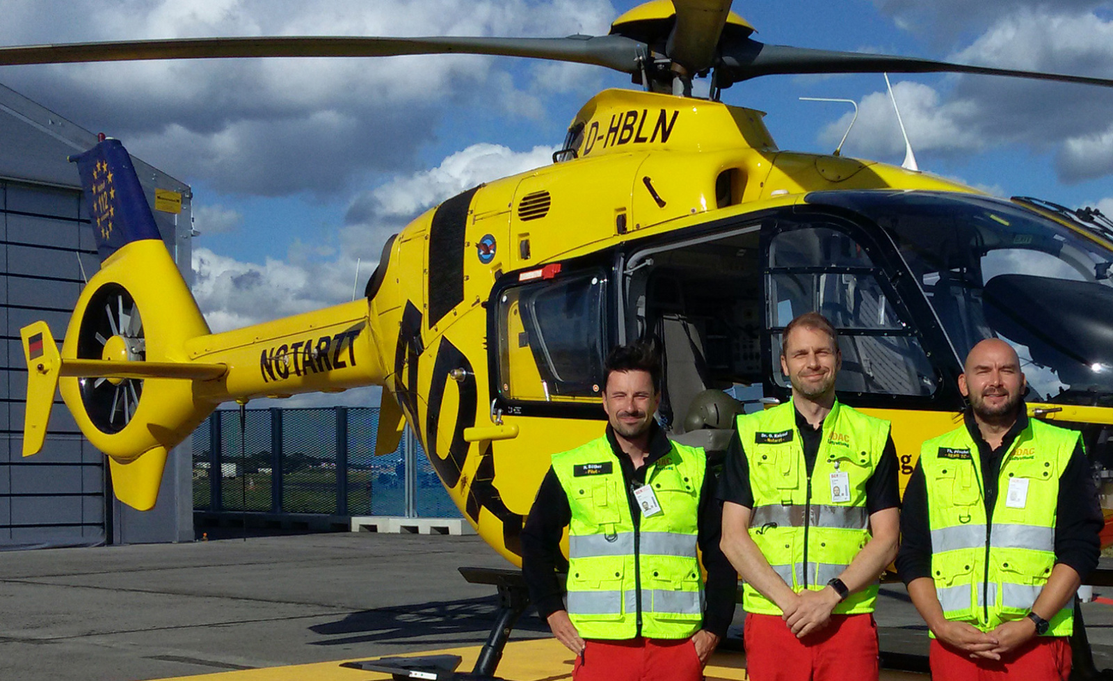 Drei ADAC Luftrettung-Teammitglieder in Warnwesten stehen vor gelbem Rettungshubschrauber mit NOTARZT-Schriftzug.