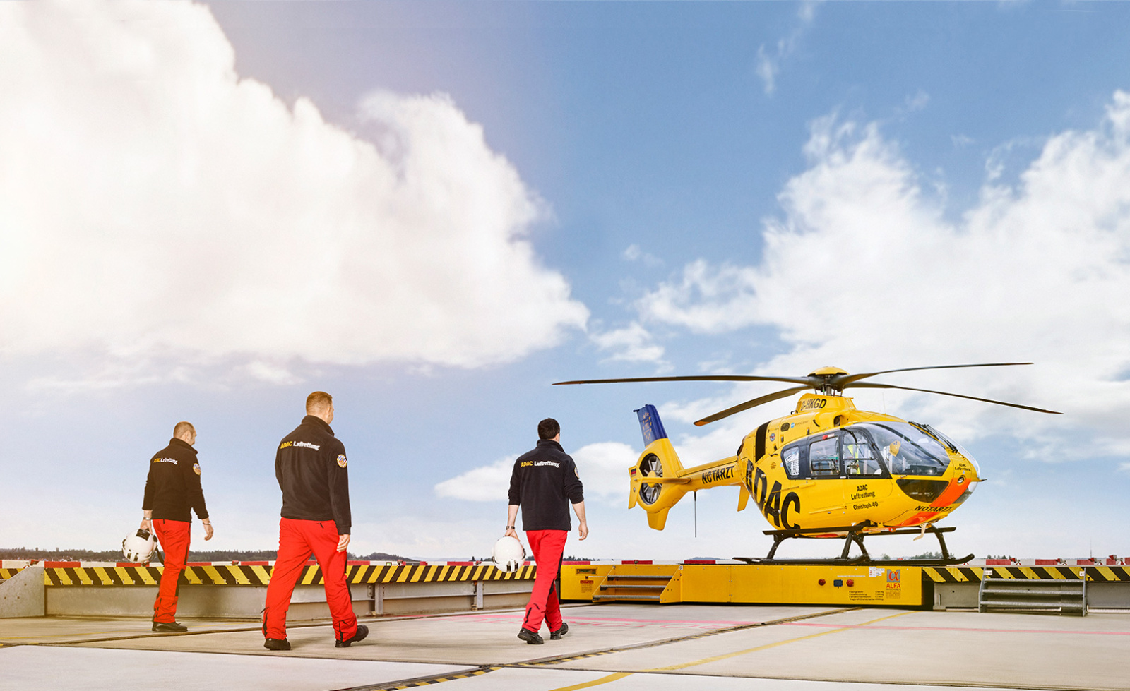 Drei ADAC Luftrettung-Crewmitglieder in schwarzen Jacken und roten Hosen gehen zum gelben Rettungshubschrauber auf Helipad.