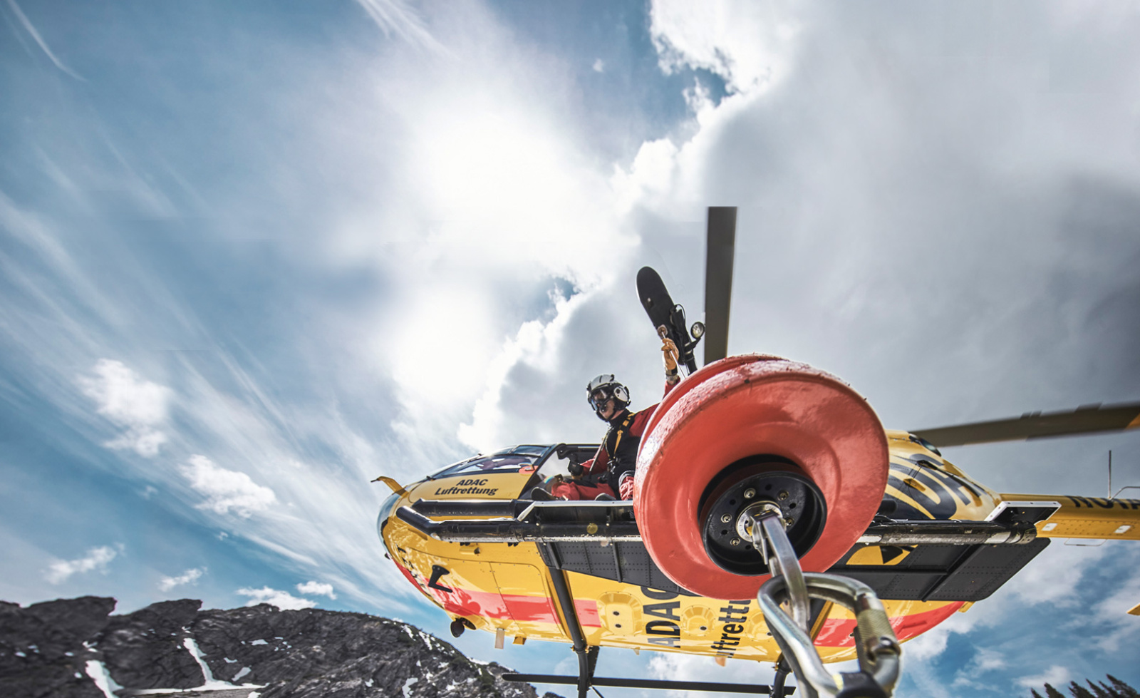 ADAC Luftrettung Rettungshubschrauber über verschneitem Gebirge, Besatzungsmitglied mit Helm am offenen Seitenausstieg.