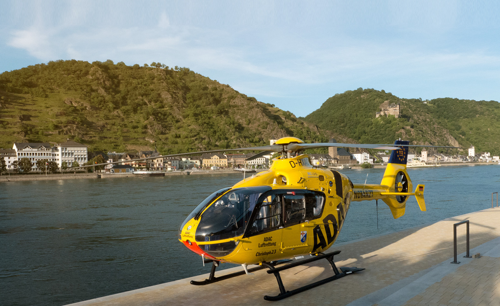 Gelber Rettungshubschrauber der ADAC Luftrettung am Flussufer, im Hintergrund Weinberge und Ortschaft
