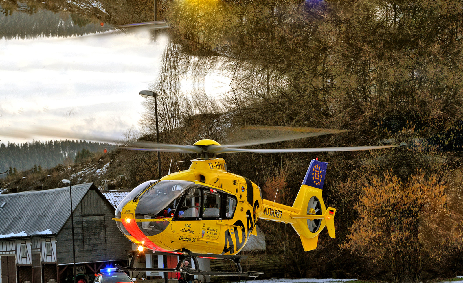 Gelber ADAC Rettungshubschrauber (ADAC Luftrettung) landet nahe Häusern, Besatzung und Einsatzwagen mit Blaulicht.