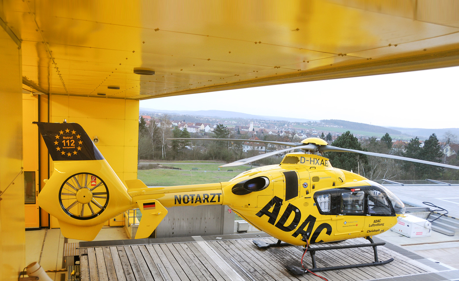 ADAC Luftrettung Rettungshubschrauber mit Notarzt-Aufschrift geparkt auf Dachlandeplatz vor offener Halle, Blick auf Stadt.
