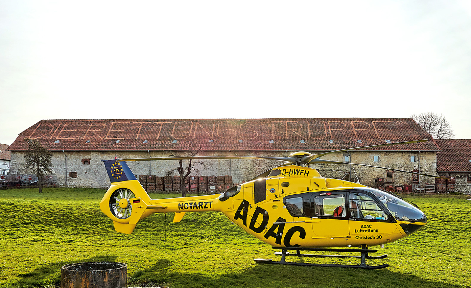 Gelber Rettungshubschrauber der ADAC Luftrettung auf Wiese vor Scheune mit Schriftzug "Rettungstruppe".