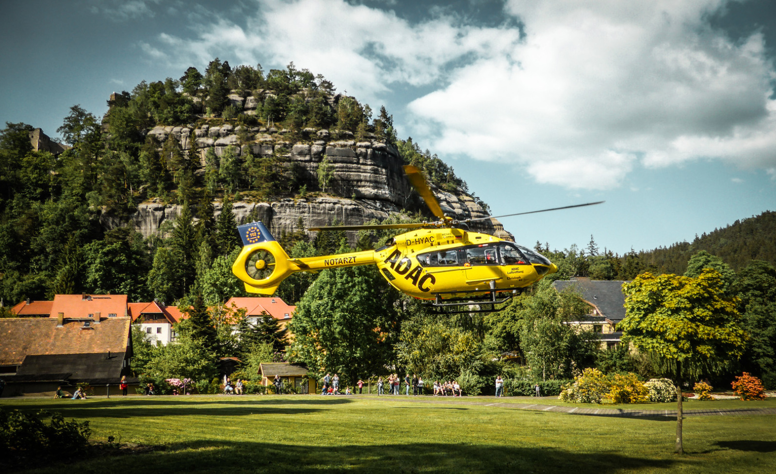 Gelber Rettungshubschrauber der ADAC Luftrettung schwebt über Park vor Felskulisse, Passanten beobachten.