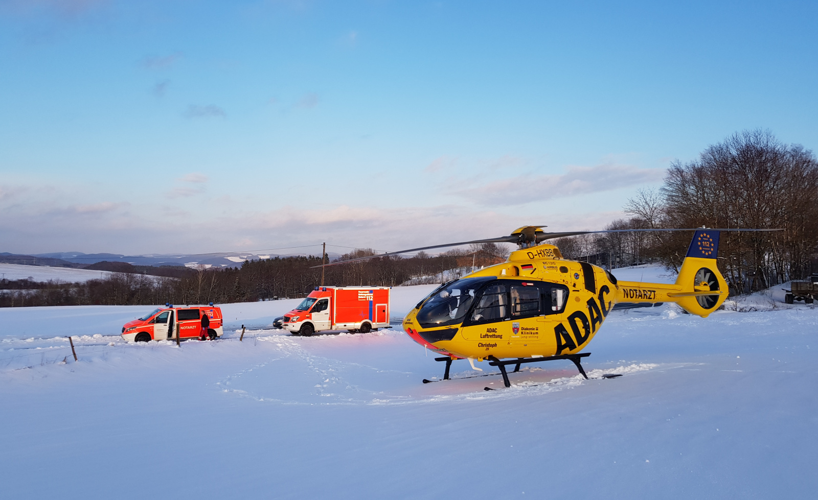 ADAC Luftrettung-Rettungshubschrauber neben Notarzt- und Rettungsfahrzeugen auf verschneiter Wiese im Einsatz