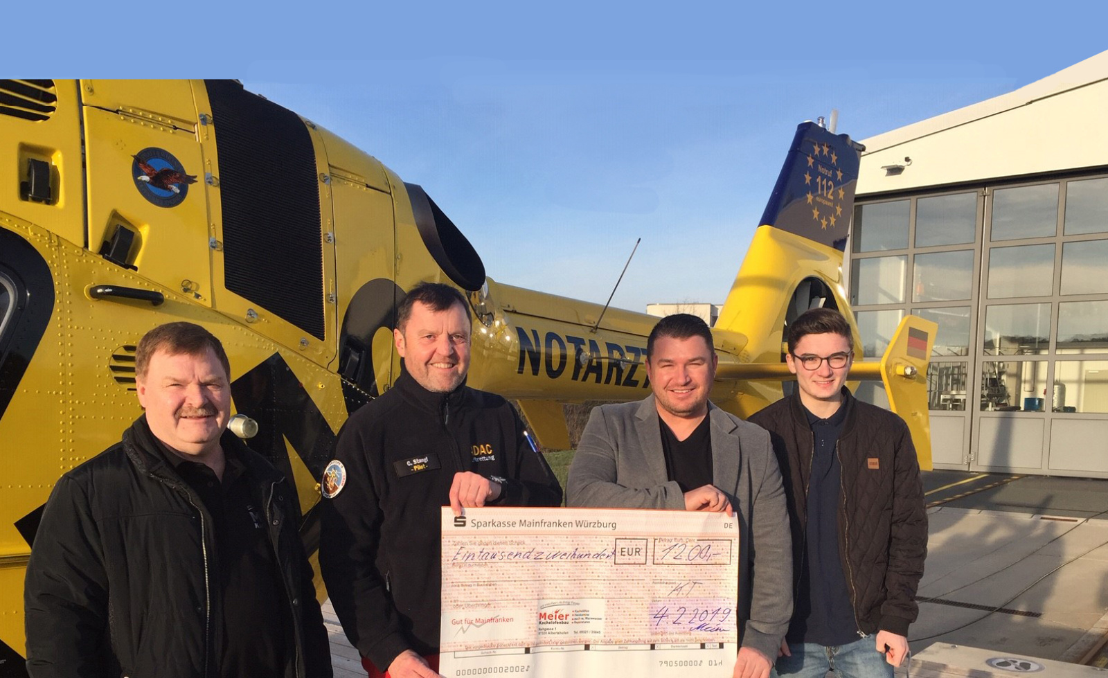 Vier Männer halten vor einem gelben Rettungshubschrauber der ADAC Luftrettung einen großen Spendenscheck.