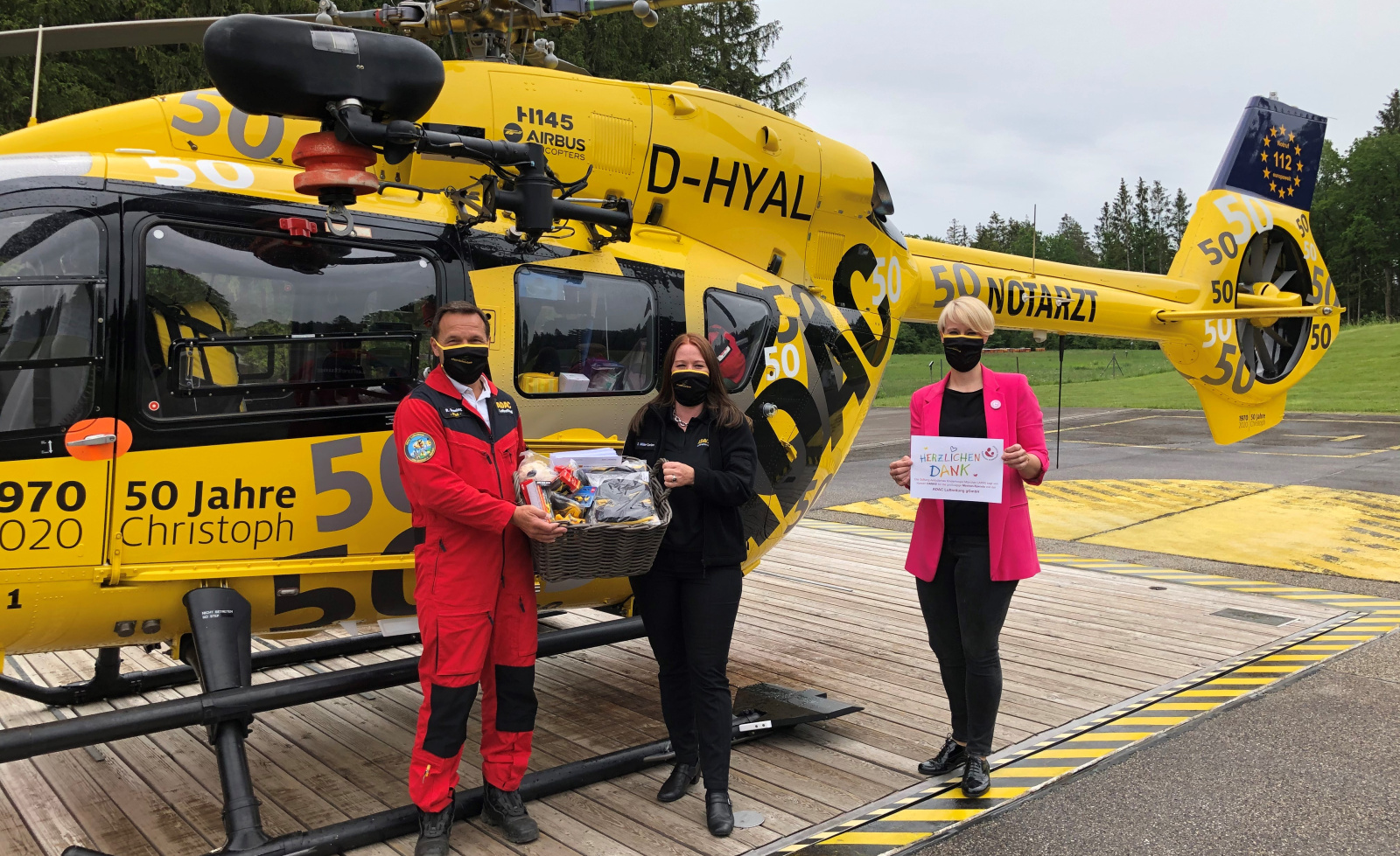 Drei Personen mit Masken übergeben einen Geschenkkorb vor dem gelben Rettungshubschrauber Christoph der ADAC Luftrettung.