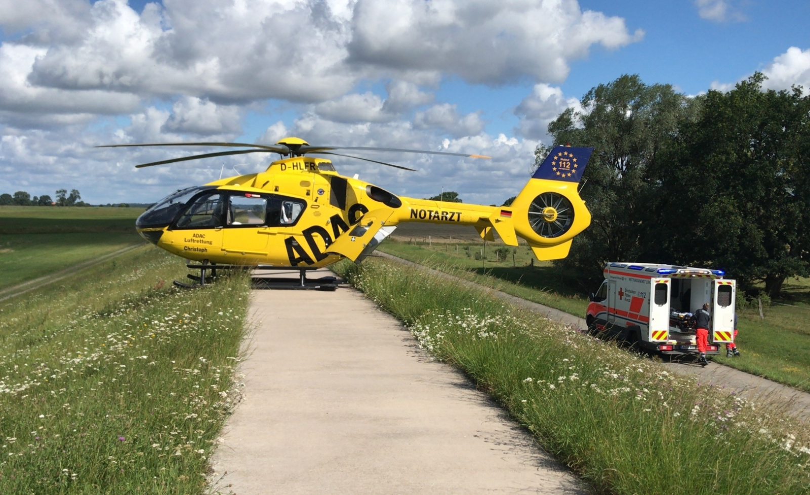 ADAC Luftrettung Rettungshubschrauber landet auf Damm, neben Rettungswagen bereitet Notarztteam Patientenübernahme vor.