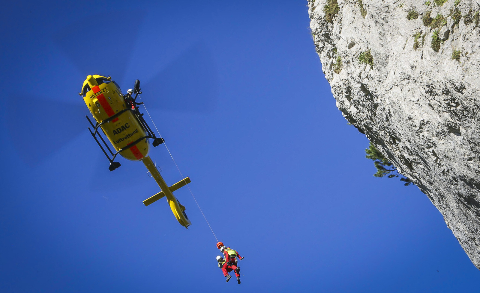 ADAC Luftrettung-Rettungshubschrauber wincht Bergretter per Seil neben steiler Felswand.