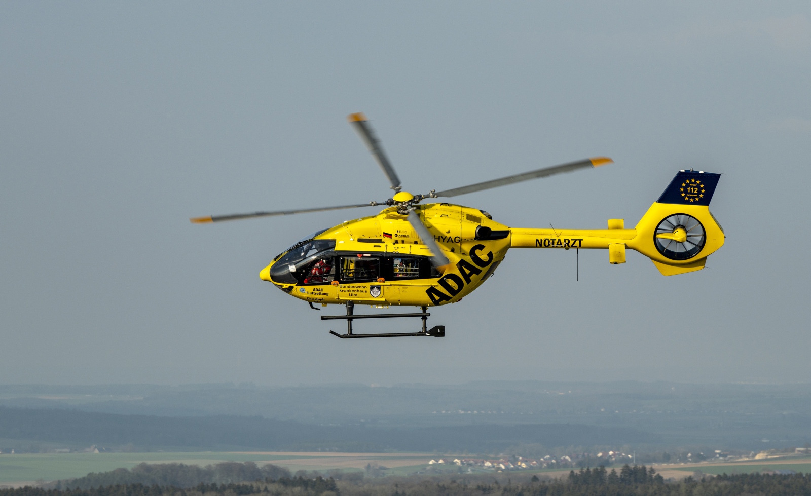 Gelber ADAC Luftrettung Rettungshubschrauber im Flug über ländliche Landschaft, Besatzung durch Kabine sichtbar.