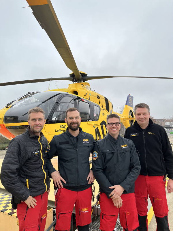 Vier Crewmitglieder der ADAC Luftrettung stehen vor gelbem Rettungshubschrauber auf Landeplatz.