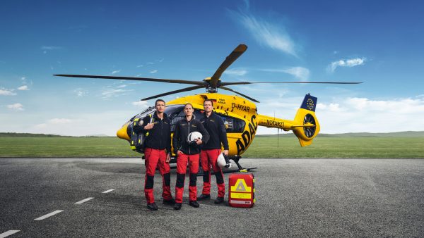 Drei Rettungsfachkräfte in Einsatzkleidung stehen vor gelbem Rettungshubschrauber der ADAC Luftrettung auf der Landebahn.